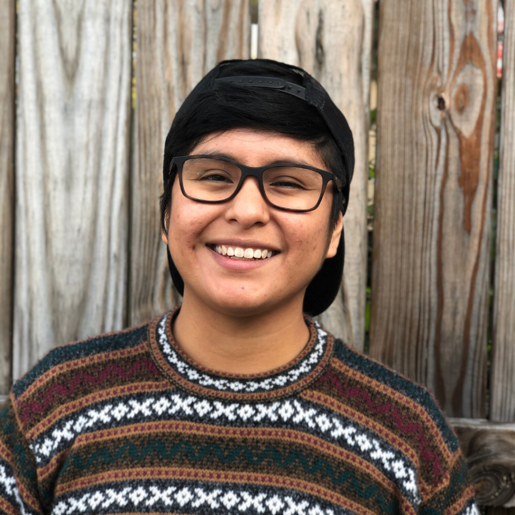 queer latinx genderqueer person in sweater smiling with glasses